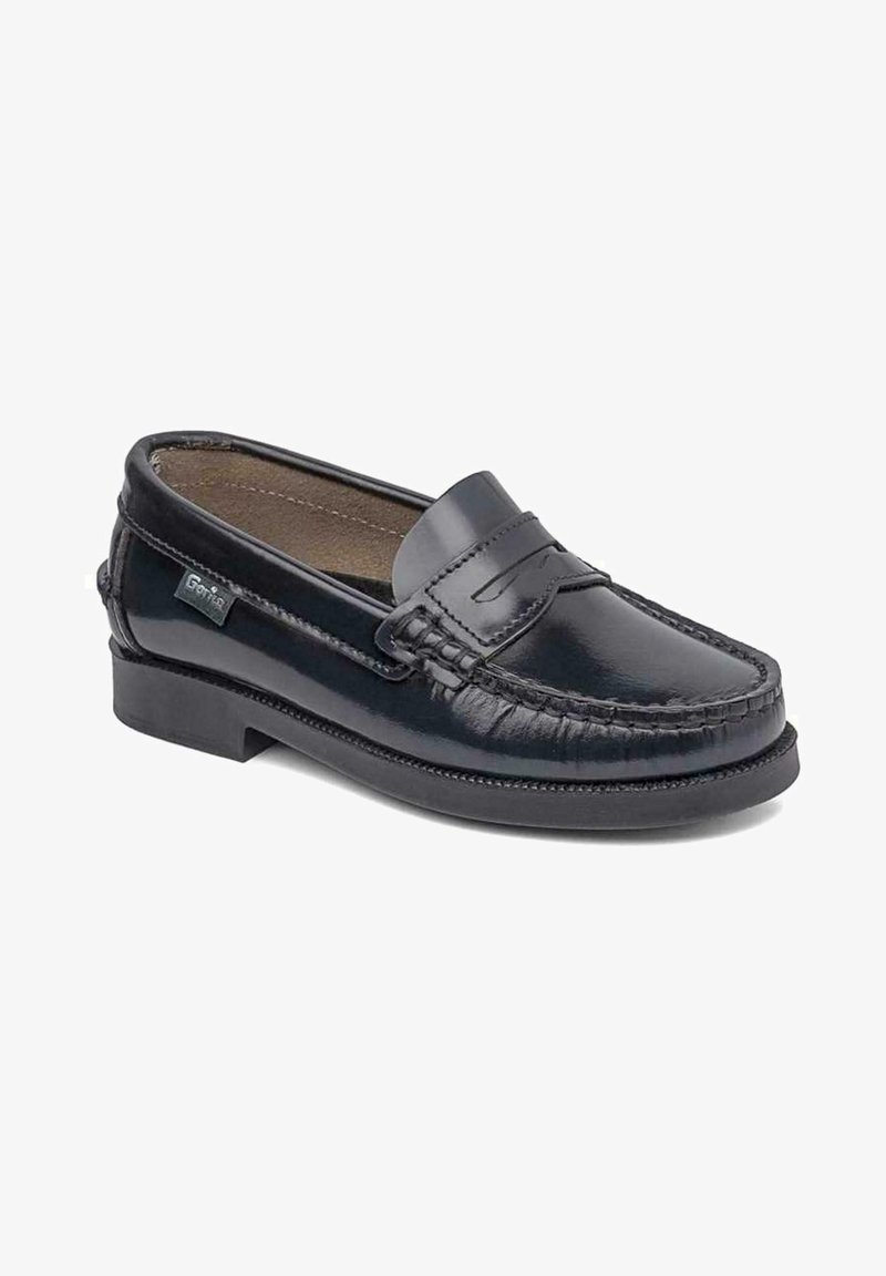 Mocasin de cuero negro con punta redondeada, costuras decorativas y un pequeño acento cosido en la parte frontal. Presenta un tacón bajo y una textura suave.