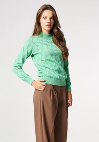 Suéter de punto verde menta con un escote alto, patrón ondulado texturizado y puños acanalados. Combinado con pantalones marrones de tiro alto.