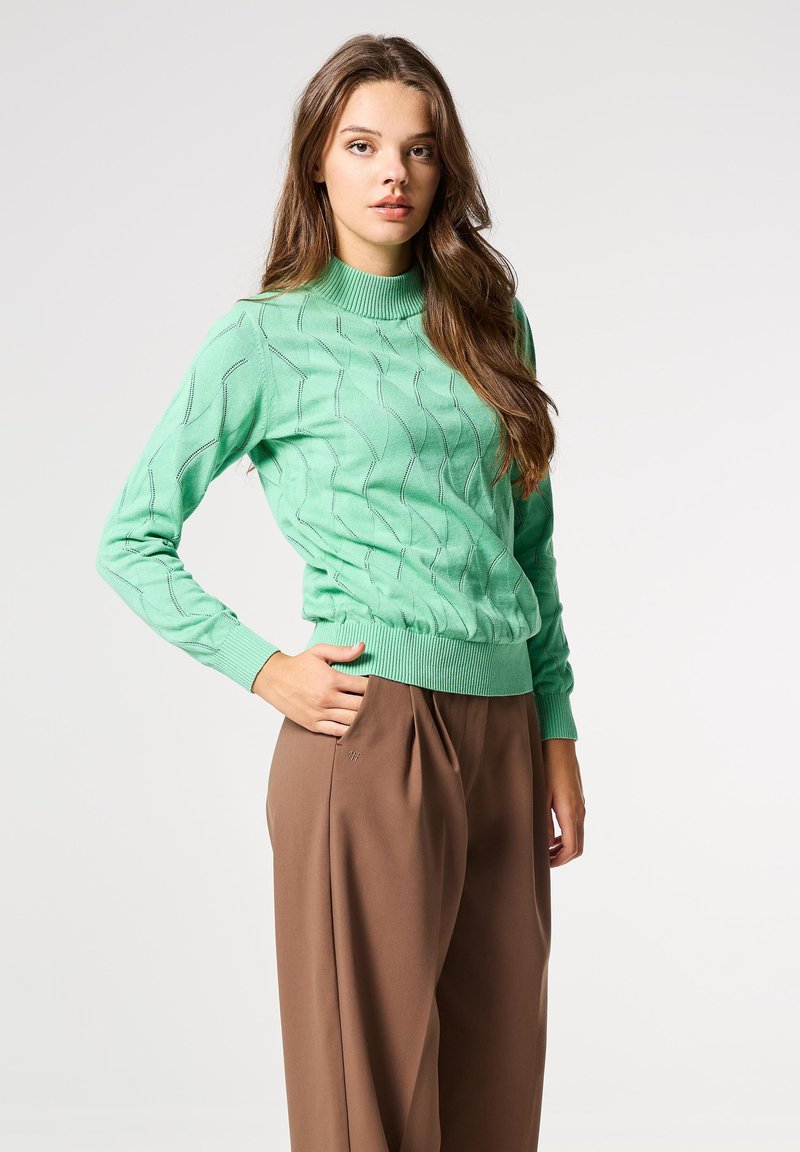 Suéter de punto verde menta con un escote alto, patrón ondulado texturizado y puños acanalados. Combinado con pantalones marrones de tiro alto.