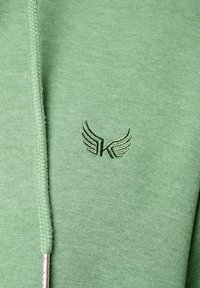 Haut de sweat à capuche vert clair en tissu doux, avec une fermeture éclair et un logo brodé d'ailes vert foncé sur la poitrine.