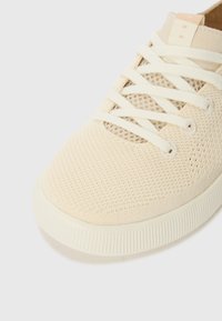 Baskets en mesh beige avec un bout rond, des lacets plats et une semelle en caoutchouc présentant un motif texturé, conçues pour la respirabilité et le confort.