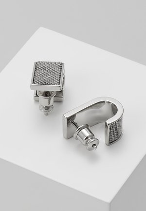 Paire de boucles d'oreilles en métal argenté avec texture en maille, une de forme carrée et une incurvée, présentées sur une surface blanche.