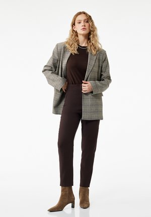 Mujer rubia con pelo ondulado que lleva una chaqueta gris de cuadros, camiseta y pantalones marrón oscuro, y botines de ante marrón, de pie con confianza.
