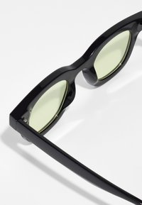 Ochelari de soare negri cu un cadru îndrăzneț, având lentile verzi și accente aurii în jurul lentilelor. Textură netedă cu elemente de design unghiular.