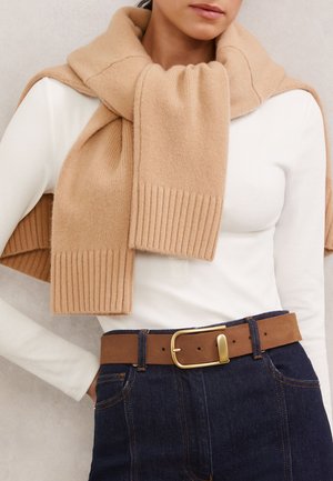 Femme portant un pull en maille beige par-dessus un top blanc à manches longues, associé à un jean bleu foncé et une ceinture marron avec une boucle dorée.