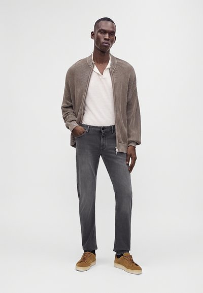 Homme portant un cardigan marron à fermeture éclair, une chemise blanche, un jean gris et des baskets beiges, se tenant devant un fond gris clair uni.