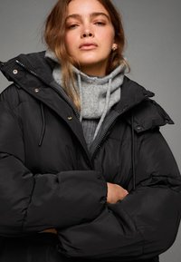 Černá péřová bunda s vysokým límcem a nastavitelnou kapucí. Má velké polstrované části a zapínání na zip, přes šedý svetr.