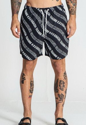 Sorte svømmebukser med hvid tekst, der gentager "GIANNI KAVANAGH." Elastisk talje med snøre. Glat stof. Tilgængelig med tatoveringer synlige på benene.