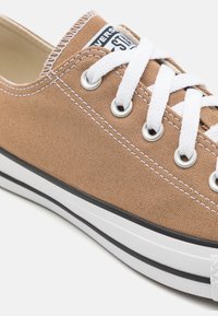 Béžové plátěné tenisky s bílými tkaničkami, kovovými očky a černým pruhovaným gumovým podešví. Mají klasický nízký design a štítek s logem.