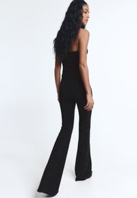 Hosszú fekete hajú nő csillogó fekete váll nélküli felsőben és bő szárú nadrágban, fehér háttér előtt áll, oldalra néz.