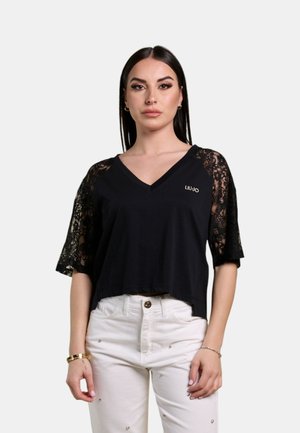 Donna con maglietta nera a scollo a V con maniche di pizzo e pantaloni bianchi con piccoli borchie color argento, in piedi davanti a uno sfondo grigio chiaro.