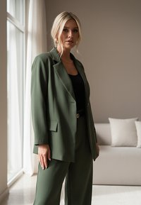 Zielona marynarka i dopasowane spodnie z lekkiego materiału. Posiadają luźny krój, duże klapy oraz przednie kieszenie. Czarna bluzka i złoty pasek w akcentach.