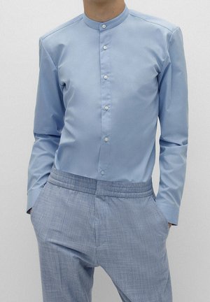 Muž nosící světle modrou košili s mandarinkovým límečkem a knoflíky, v kombinaci se světle modrými texturovanými kalhotami, ruce v kapsách.