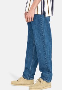 Jeans in denim blu con vestibilità rilassata e cuciture visibili, abbinati a scarpe in suede color ocra. Il tessuto è testurizzato e leggermente sbiadito.