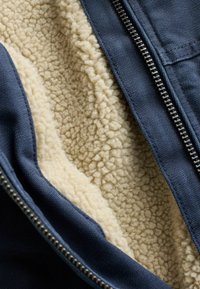 Veste bleu marine avec fermeture éclair argentée partiellement ouverte pour révéler la doublure polaire douce beige à l'intérieur.