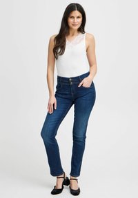 Bílé tílko s krajkovým výstřihom spárované s tmavomodrými skinny džíny a černými sandály na podpatku, stojící na světlém pozadí.