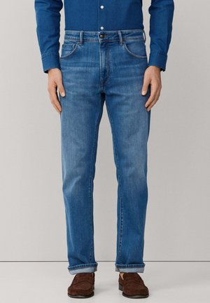 Man draagt blauwe denim jeans met opgerolde pijpen, bruine suède loafers en een blauw overhemd, staand tegen een effen achtergrond.