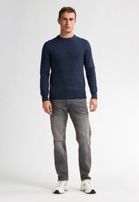 Marineblauwe gebreide trui met een gestructureerd patroon, ronde hals, en geribde mouwen, gecombineerd met grijze jeans en witte sneakers.