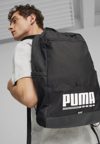 Juodas kuprinė, pagaminta iš tvirto audinio, su priekiniais užtrauktukais ir tinkliniais šoniniais kišenėmis. Didelis baltas logotipas ir tekstas priekyje.