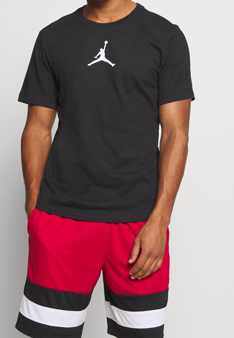 Man som bär en svart Jordan T-shirt med vitt Jumpman-logotyp och shorts i blockfärger i rött, svart och vitt, stående mot en enfärgad bakgrund.