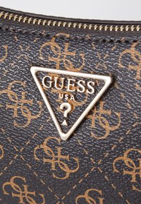 Materiale nero testurizzato con un motivo a logo marrone, con una medaglia triangolare dorata che riporta "GUESS USA" e un punto interrogativo.