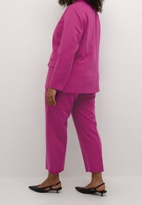 Mujer vestida con un traje magenta que incluye pantalones recortados y una blazer, combinado con zapatos bajos de tacón en la parte trasera, de pie de perfil.