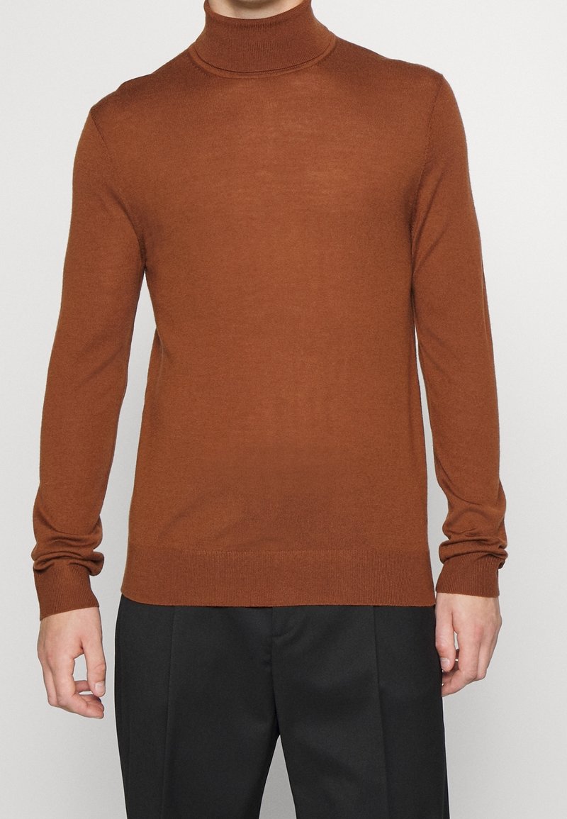 Mann, der einen enganliegenden braunen Rollkragenpullover und schwarze Hose trägt und vor einem schlichten hellen Hintergrund steht.