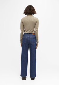 Jean en denim bleu à jambe large avec une taille haute, présentant une patch en cuir marron à l'arrière et des coutures détaillées. Association avec un haut beige ajusté.
