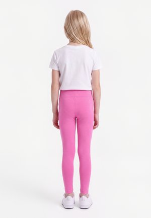 Jeune fille aux longs cheveux blonds portant un t-shirt blanc à manches courtes, des leggings roses et des baskets blanches, debout de dos.