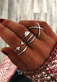 Silver rings displayed on a hand, featuring various designs: a sunburst, a braided pattern, a cross, and flower shapes with gemstones.
