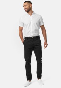Camisa blanca de botones de manga corta, pantalones negros y zapatillas blancas sin cordones. Tejido de algodón suave, ajuste casual y detalles sutiles en los pantalones.