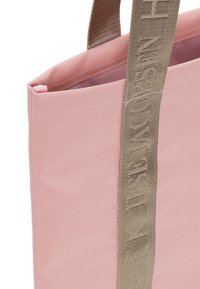 Pink tote bag made of durable fabric with a textured finish. Features a wide beige strap with embossed lettering along the side.