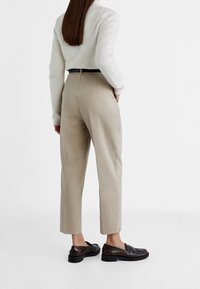 Beige, gepaste broek met een rechte pijp en een lichte taper, gecombineerd met een lichtgrijze, fluffy trui. Bruine instappers maken de outfit compleet.