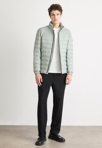 Light green quilted puffer jacket with a high collar and zip closure, paired with black trousers and black shoes on a neutral background.