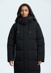 Manteau doudoune noir avec un col haut, des poches zippées, des accents argentés, un design matelassé et des cordons ajustables sur la capuche.