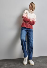 Maglione a maglia grossa nei colori crema, rosa e rosso a righe; design con colletto alto. Abbinato a jeans a gamba larga blu e sneakers bianche.