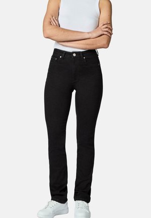 Person iført tætsiddende sorte jeans, hvid ærmeløs top, hvide sneakers, stående med arme krydset mod en ensfarvet baggrund.