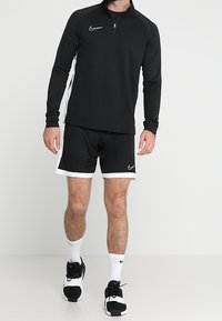 Svart atletisk tröja med kvartdragkedja och vita detaljer, parat med svarta shorts med vita kanter. Bärs med svarta sneakers och vita strumpor.