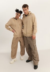 Felpe beige con zip e coulisse, pantaloni cargo color sabbia, pantaloni a quadri marroni e sneakers a contrasto, esposti su uno sfondo neutro.