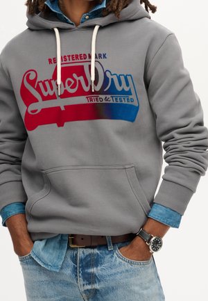 Mężczyzna ubrany w szarą bluzę z kapturem Superdry z czerwononiebieskim logo, pod spodem koszula dżinsowa, jasnoniebieskie dżinsy, brązowy pasek i czarny zegarek na nadgarstku.