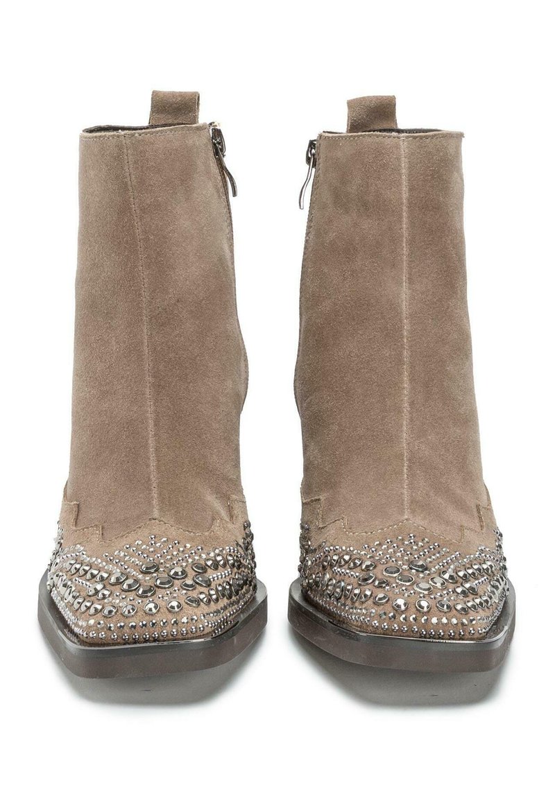 Botines de ante color tostado con punta afilada, que presentan tachuelas decorativas de plata y bordes en forma de ondas. Cierre lateral con cremallera para facilitar su uso.