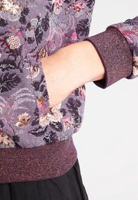 Bomberka w kwiatowy wzór z fioletowego materiału, z złotymi dodatkami, prążkowanymi mankietami i boczną kieszenią, o luźnym kroju.