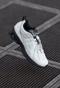 Scarpa sportiva bianca con accenti neri. Presenta una tomaia in mesh testurizzato, suola ammortizzata e design con lacci. Logo "N" ben visibile sul lato.