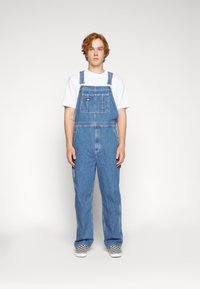 Világoskék farmer overall állítható pántokkal, elülső zsebbel, széles lábakkal, fehér pólóval és kockás slip-on cipővel párosítva.