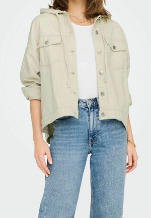 Femme portant une veste boutonnée beige clair sur une chemise blanche et un jean bleu taille haute, avec les manches retroussées et des bijoux en or au poignet et au cou.