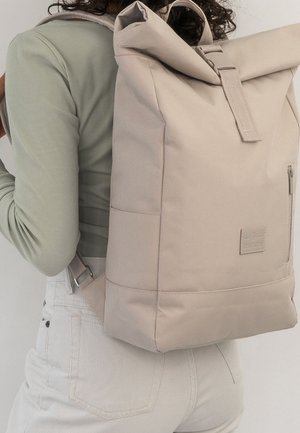 Persoon met een lichtgroene top met lange mouwen en een witte broek, draagt een grote beige roll-top rugzak met voorvak met rits.