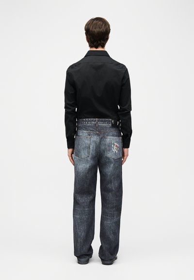 Άνδρας πλάγια με μαύρο πουκάμισο μακρυμάνικο και χοντρό γκρι τζιν με ένα μικρό καρτούν σχέδιο στην δεξιά πίσω τσέπη.