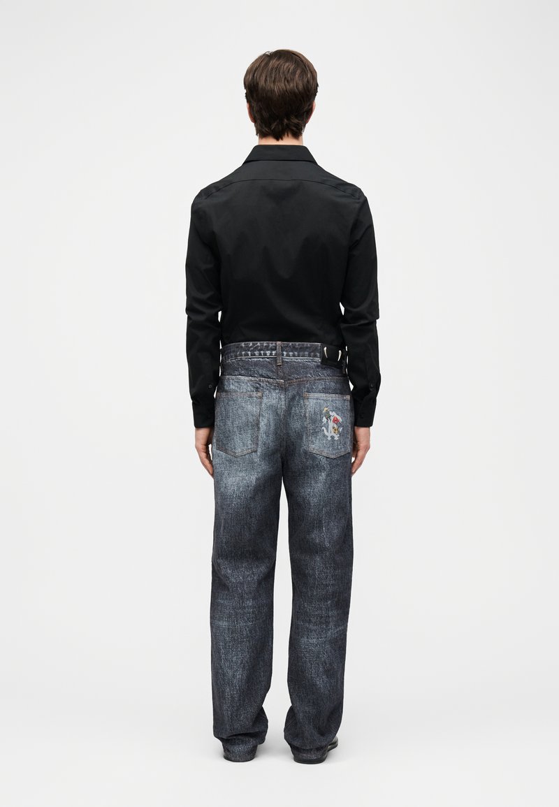 Homme de dos portant une chemise noire à manches longues et un jean gris ample avec un petit patch cartoon sur la poche arrière droite.