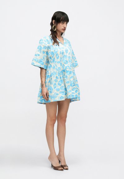 Γυναίκα με σκούρα μαλλιά που φορε ί μεγάλα χρυσά σκουλαρίκια κρίκους, κοντό φόρεμα με μπλε και λευκά σχέδια και καφέ ψηλοτάκουνα παπούτσια, στέκεται μπροστά σε λευκό φόντο.