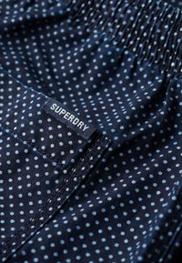 Shorts bleu marine avec des pois bleu clair, taille élastique et une étiquette visible portant "SUPERDRY" sur la poche latérale.